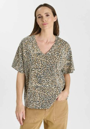 Donna che indossa una blusa a maniche corte con scollo a V e stampa leopardata con pantaloni beige, in piedi con una mano in tasca su uno sfondo semplice.