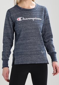 Donna che indossa una felpa blu Champion con il logo sul petto e leggings neri, in piedi contro uno sfondo chiaro e semplice.