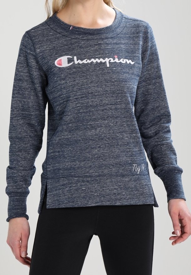 Donna che indossa una felpa blu Champion con il logo sul petto e leggings neri, in piedi contro uno sfondo chiaro e semplice.