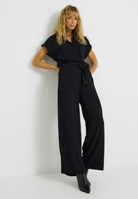 Combinaison noire en tissu texturé, avec des manches courtes, un devant boutonné et une ceinture à nouer à la taille. Design à jambes larges.