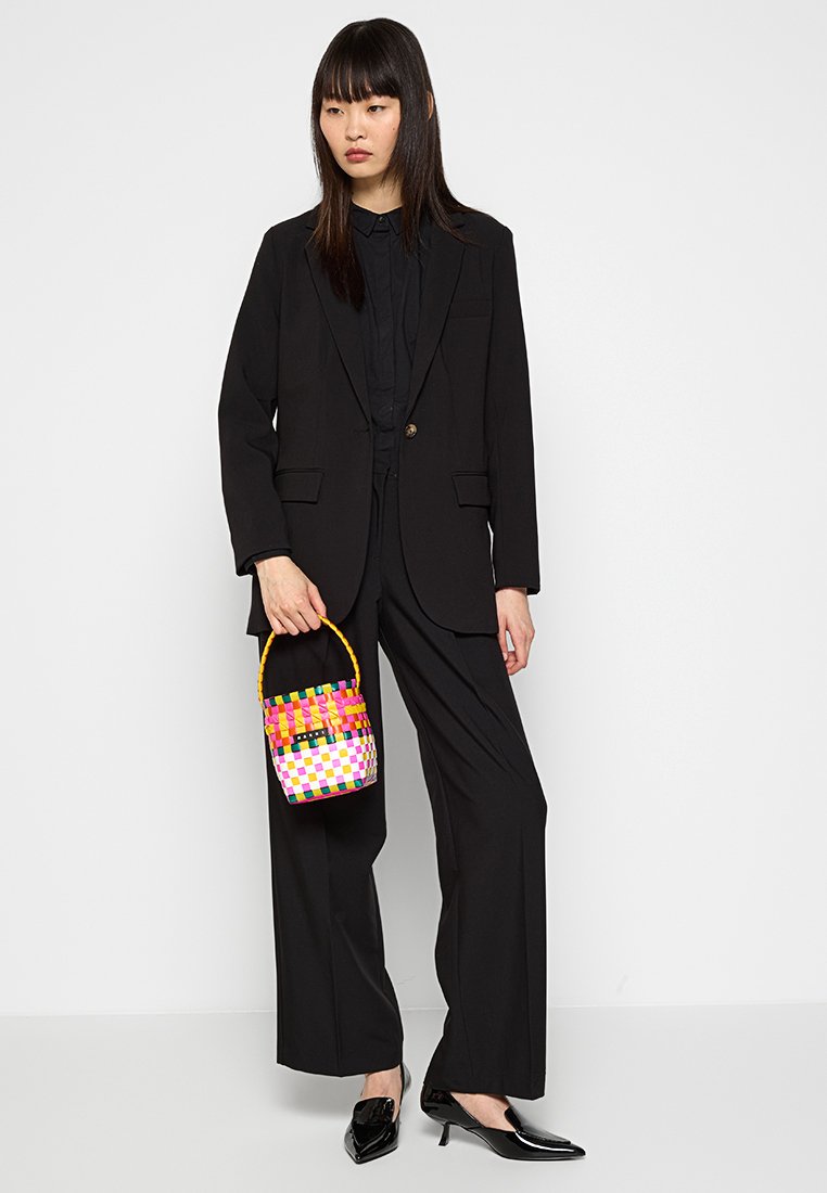 Schwarzer taillierter Anzug mit einer taillierten Blazerjacke und weiten Hosen. Das Model hält eine bunte geflochtene Bucket Bag mit einem gelben Griff. Schwarze Lackschuhe.