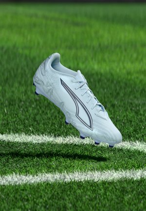 Bota de fútbol blanca con logo negro flotando sobre el campo de césped verde cerca de una línea límite blanca.