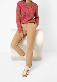 Pull en tricot beige avec motif abstrait rose ; associé à un pantalon beige et des baskets blanches avec des accents et des lacets bruns.
