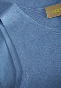 Niebieski sweter dziewiarski z dekoltem w kształcie litery V, prążkowanymi akcentami i gładką fakturą. Metka z napisem "JJXX" w żółtym na jasnobrązowym tle.