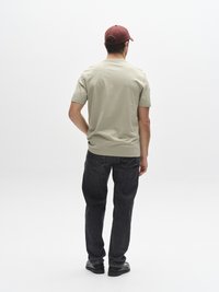 Beige kortärmad t-shirt med rund hals, kombinerad med mörka denimjeans. Mörkröd keps, svarta skor, sedd bakifrån mot en enfärgad bakgrund.