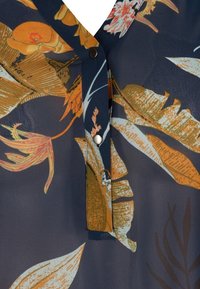 Blouse marine ornée de motifs floraux vibrants en orange, jaune et bleu. Elle possède une patte de boutonnage et une texture douce et transparente.