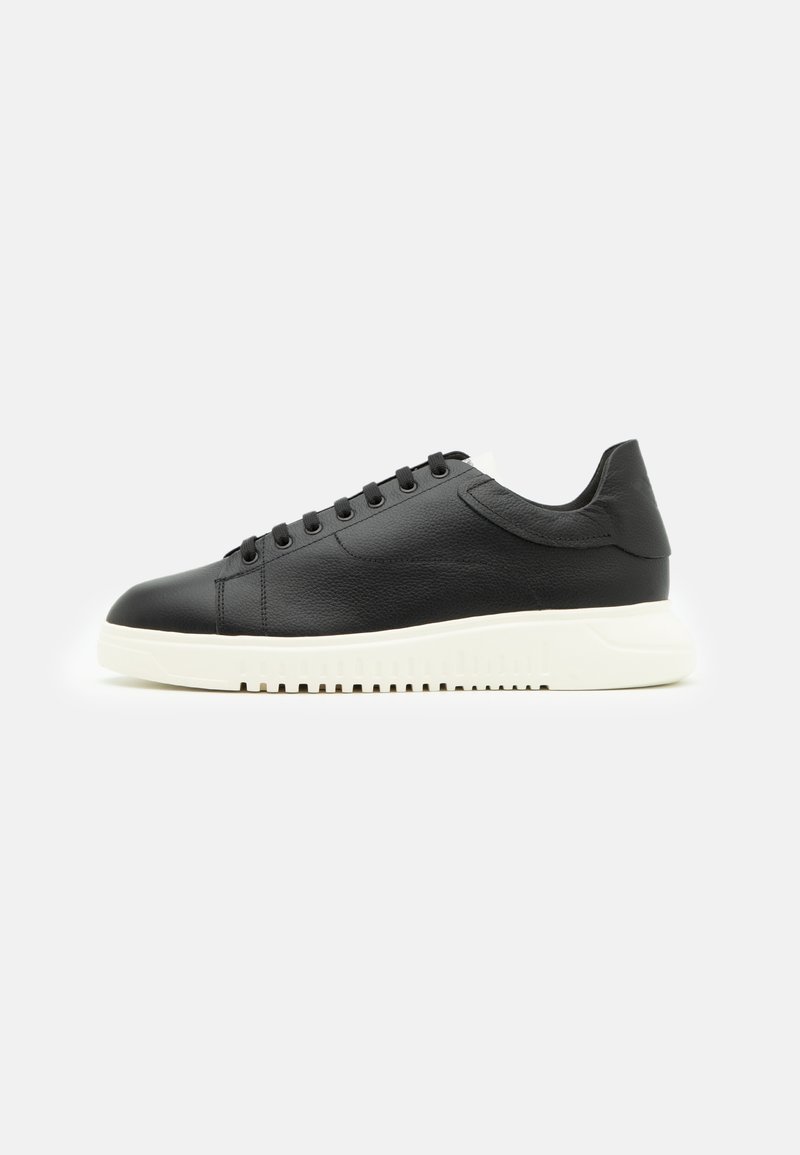 Schwarzer Low-Top Sneaker mit schwarzen Schnürsenkeln und strukturiertem Lederobermaterial, mit einer dicken weißen profilierten Sohle auf hellem Hintergrund.