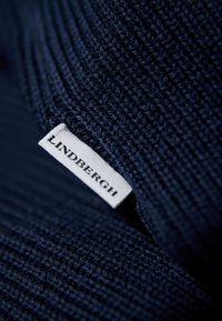 Marinblått stickat tyg med texturerad yta, med en vit etikett med texten "LINDBERGH" fäst vid kanten.