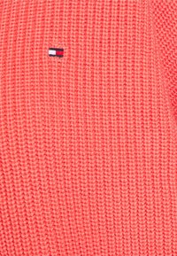 Korallfärgad stickad tröja med en strukturerad yta. Har ett litet emblem i mörk marinblått, rött och vitt nära bröstet.