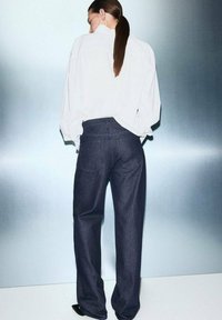 Jeans de mezclilla oscuros de corte holgado y tiro alto, combinados con una camisa blanca de gran tamaño, vistos desde la parte de atrás contra un fondo degradado.
