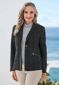 Veste tissée noire avec des accents métalliques, revers crantés et poches zippées. Stylée sur un haut gris clair, associée à un pantalon crème.