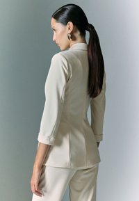 Blazer fitted beige con maniche a tre quarti, design strutturato e leggero lucido. Abbinato a pantaloni su misura coordinati, texture liscia.