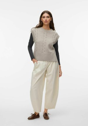 Femme portant un pull sans manches gris torsadé par-dessus des manches longues noires, un pantalon beige ample et des chaussures marron à lacets, debout avec une main dans la poche.