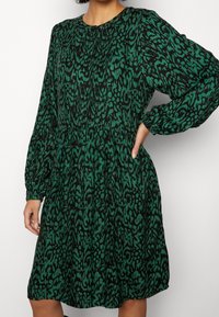 Vestido de patrón verde y negro, de mangas largas, con cuello redondo, cintura fruncida y falda ligeramente acampanada, que presenta un sutil diseño de estampado animal.