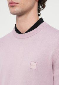 Svetlo vijoličen pulover z okroglim izrezom in črnim ovratnikom, z majhnim kvadratnim logotipom v svetlo rožnati barvi na prsnem delu.