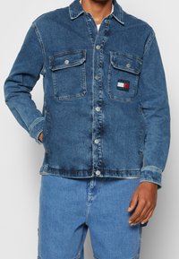 Giacca di denim blu scuro con chiusura a bottoni, due tasche sul petto e una piccola etichetta con il logo. Presenta un collo a camicia e una texture leggermente sbiadita.