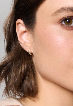 Złota kolczyk w kształcie obręczy z przezroczystymi kamieniami, z zakrzywionym wzorem. Zbliżenie ukazuje gładką powierzchnię i subtelny blask.