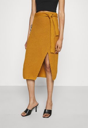Gebreide mosterdgele midirok met voorsplit en ceintuur, gedragen met een zwarte mouwloze top en zwarte open-teen sandalen met hak.