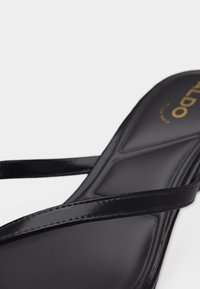 Musta sandaali, jossa on kaksi ohutta kiiltävää hihnaa pehmustetun jalkapohjan päällä ja joka paljastaa osittain kullanvärisen tuotemerkin logon kantapään kohdalla.