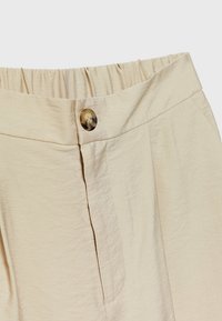 Pantalon beige avec ceinture élastique et un bouton en écaille à la fermeture avant, montrant la texture du tissu et les détails des coutures.