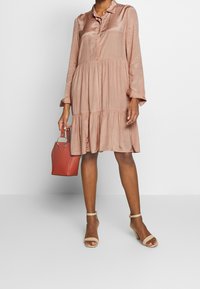 Donna che indossa un vestito lungo a maniche lunghe, morbido e lucido, di un rosa polveroso, con sandali con tacco beige, mentre tiene una piccola borsa rossa con manici ad anello in metallo.