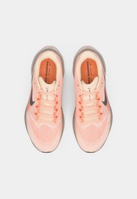Une paire de chaussures de course Nike corail clair avec des logos swoosh noirs, des lacets roses et des semelles intérieures orange, vues de dessus sur un fond blanc.