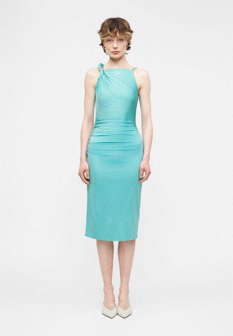 Γυναίκα που φοράει στενό, αμάνικο τουρκουάζ φόρεμα μήκους μέσης με δέσιμο στον ώμο και άσπρες μυτερές γόβες, στέκεται μπροστά σε λευκό φόντο.