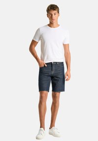 Weißes Baumwoll-T-Shirt für Herren, kombiniert mit dunklen Denim-Shorts. Die Shorts haben einen geraden Schnitt und enden knapp über dem Knie, ergänzt durch weiße Sneakers.