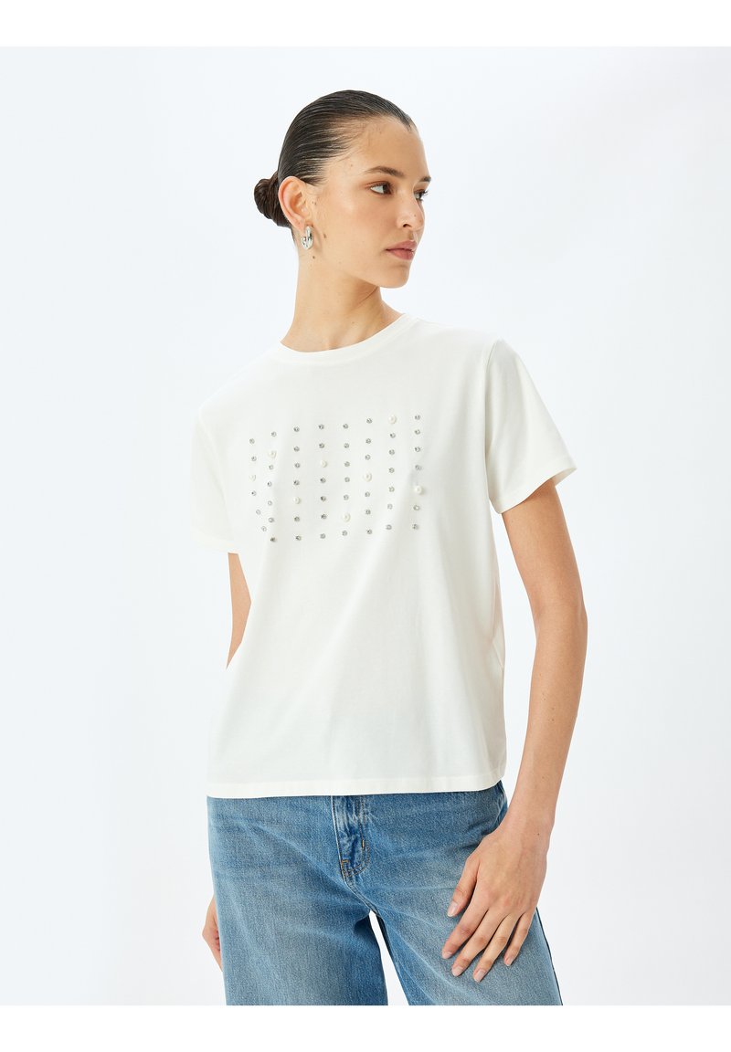 Valkoinen puuvillapaita lyhyillä hihoilla, jossa on edessä metallisten niittien ruudukko. Yhdistetty vaaleansinisiin farkkuihin.