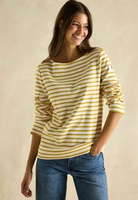 Gelb-weiße gestreifte Langarmbluse mit weitem Ausschnitt und hochgekrempelten Ärmeln, kombiniert mit blauen Jeans. Hat ein kleines Etikett am Ärmel.