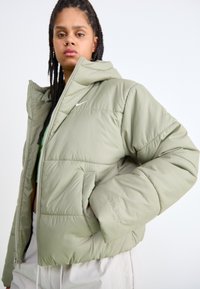 Giacca a vento verde con cappuccio, dotata di chiusura con zip, tasche laterali e imbottitura testurizzata. Logo Nike sul petto e la frase "THERMA FIT" sulla manica.