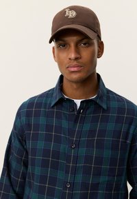 Πουκάμισο καρό σε αποχρώσεις του ναυτικού μπλε και πράσινου με κίτρινα στοιχεία και κουμπιά, συνδυασμένο με καφέ καπέλο που φέρει λογότυπο.