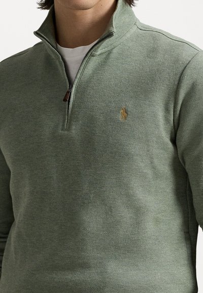 Green knit pullover with a zippered collar, featuring a small gold emblem on the chest and a textured fabric.
