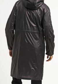 Imperméable noir à capuche avec une texture lisse et brillante, détail de taille élastique. Manches longues et coupe mi-longue. Aucun motif ou accent visible.