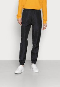 Černé joggingové kalhoty s elastickým pasem a manžetami, v kombinaci se žlutým svetrem a bílými teniskami. Hladký materiál, ležérní design.