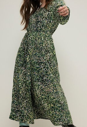 Femme portant une longue robe verte à motifs avec des manches longues, tendant un bras vers l’avant, sur un fond clair uni.
