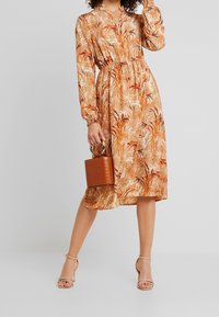 Robe beige à motifs floraux orange avec manches longues et taille cintrée. Associée à un sac à main marron texturé et à des talons à brides couleur sable.