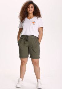 T-shirt bianco a V con grafica "Sii te stesso", abbinato a pantaloni corti verdi oliva, di lunghezza al ginocchio, con due tasche frontali e vita con cordino.
