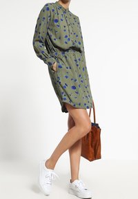 Olivově zelené šaty s květinovým potiskem v modré. Dlouhé rukávy, límeček, elastický pas. Kombinované s bílými teniskami a hnědou kabelkou.