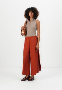Beige ribbed sleeveless top with button placket, paired with wide-legged rust pants. Brown loafers with gold hardware, holding a small brown bag.