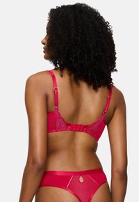 Γυναίκα με σγουρά μαλλιά που φοράει κόκκινο δαντελένιο σουτιέν και ταιριαστά εσώρουχα, φαίνεται από πίσω σε έναν απλό φωτεινό φόντο.