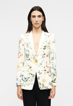 Γυναίκα με ίσια σκοτεινά μαλλιά, που φοράει ένα λευκό μπλεϊζερ με λουλούδια και πεταλούδες και μαύρο παντελόνι, στέκεται μπροστά σε απλό φωτεινό φόντο.