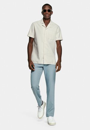 Homme portant des lunettes de soleil, une chemise beige à manches courtes, un pantalon bleu clair et des baskets blanches, marchant vers l'avant sur un fond uni.