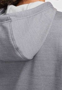 Gros plan d'un sweat à capuche gris porté par une personne aux cheveux bouclés, mettant l'accent sur la capuche et la zone des épaules.
