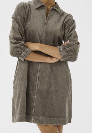 Kvinde iført en gråbrun fløjlsbuksedragt med lange ærmer, krave og en lynlås foran, armene krydset over brystet.