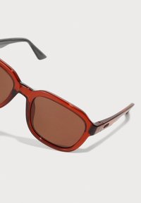 Gafas de sol transparentes rojas con montura cuadrada y lentes de color marrón; presentan acentos negros en las patillas.