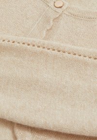 Pulover tricotat într-o nuanță moale de bej, cu fire metalizate. Are un guler rotund și detalii cu nasturi. Textură netedă cu un luciu subtil.