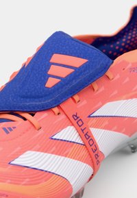 Botín de fútbol de color rosa brillante con una parte superior azul y tres franjas naranjas. Superficie texturizada con detalles en blanco y un diseño de ajuste ceñido.