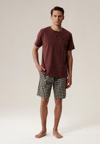 Maroonowa koszulka henley z krótkim rękawem i trzema guzikami. Szorty w zielono-białą kratkę z troczkiem. Model stoi boso.