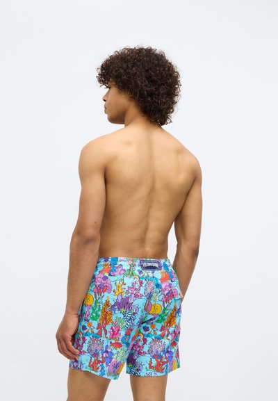 Bărbat cu păr creț stă fără tricou, întors cu spatele, purtând o pereche de șorturi de baie albastre strălucitoare cu un model colorat de recif de corali.